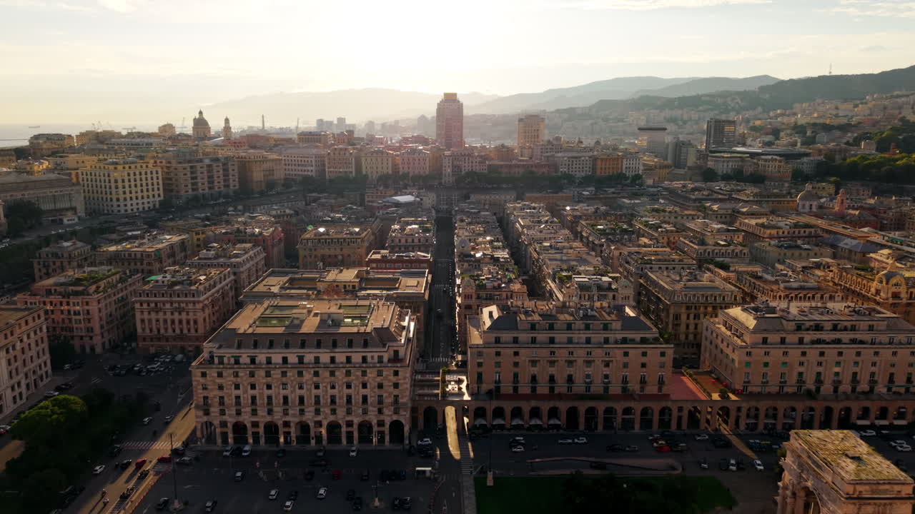 Ripresa frontale con drone che attraversa Piazza della Vittoria, mostrando l'arco trionfale, edifici simmetrici, tetti e montagne in lontananza al tramonto.
