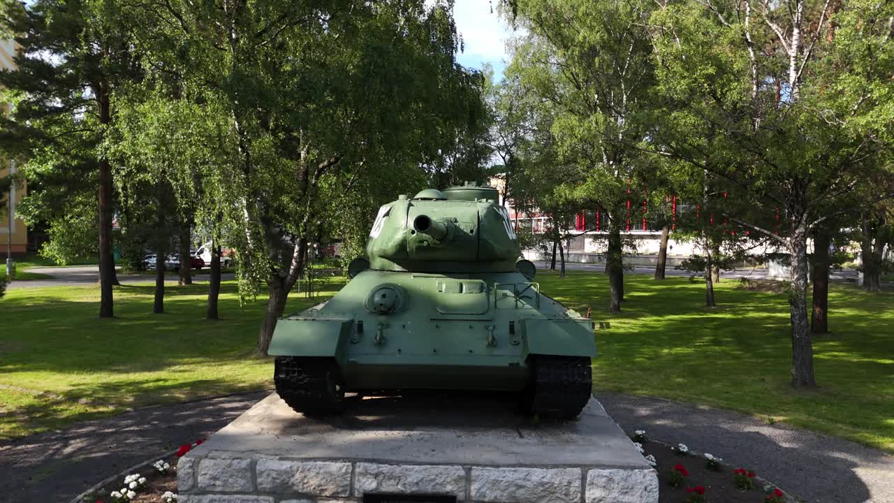 Aerial view of a T34 tank monument in a quiet park in Slovakia, surrounded by trees, grass and pathways. The preserved historic military vehicle stands on a stone pedestal in soft daylight