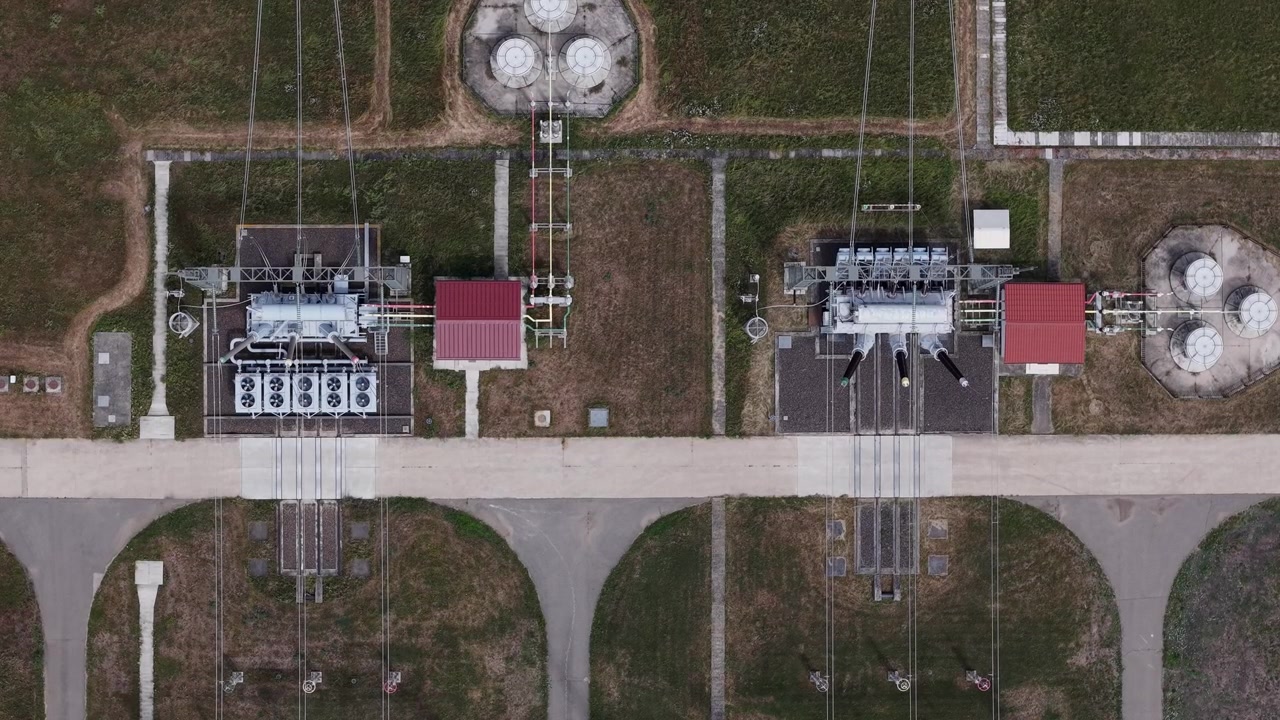 Letecký pohled z ptačí perspektivy na elektrickou rozvodnu s vysokonapěťovými transformátory distribuujícími energii napříč venkovskou krajinou.