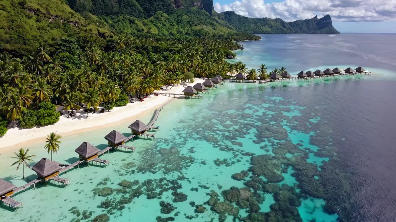 Luftvideooptagelser af bungalows over vandet på en tropisk ø, der viser turkisblåt vand og frodig vegetation