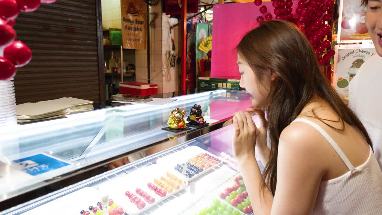 Two people select colorful tanghulu skewers from a brightly lit street food display at night