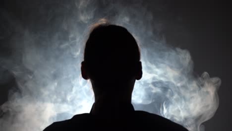 isolated silhouetted closeup of a person with long hair binge smoking in a dark room facing a dark wall