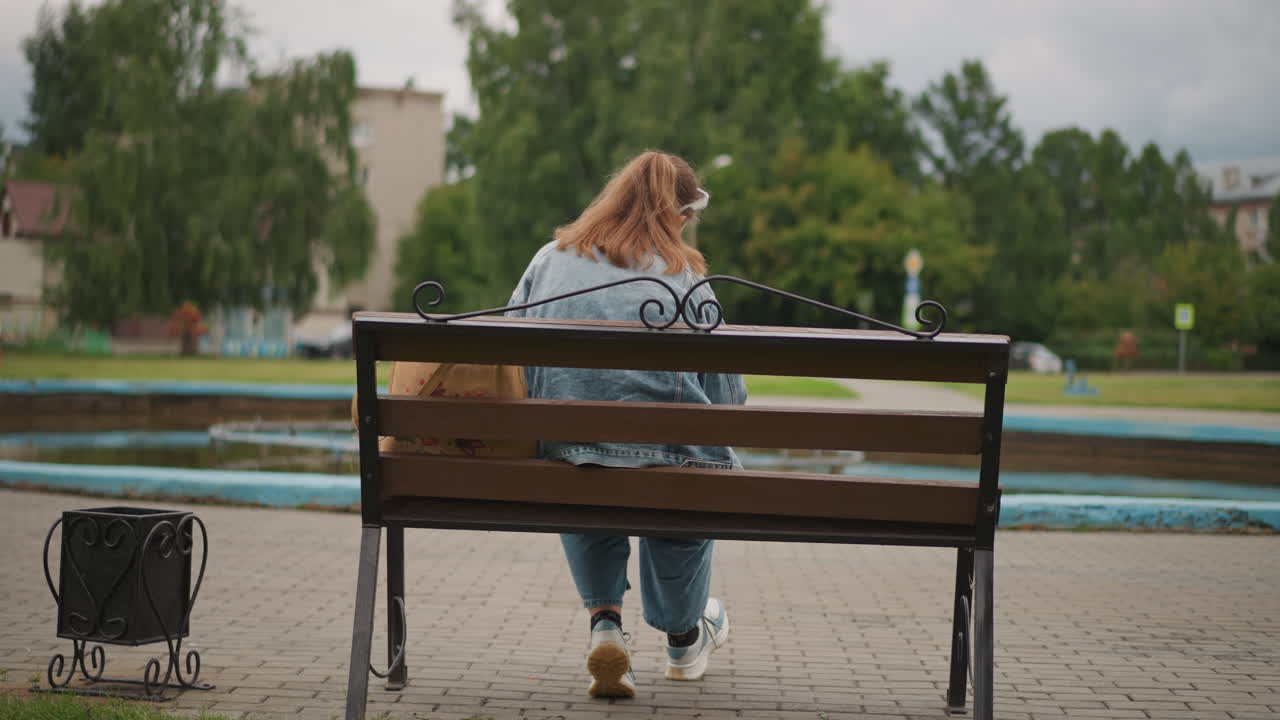 Белая женщина сидит на скамейке лицом к фонтану, в джинсовой куртке, маленькая сумка и багаж на земле, деревья и водный фон, задумчивая поза, поэтическое спокойствие, мягкое пасмурное освещение.