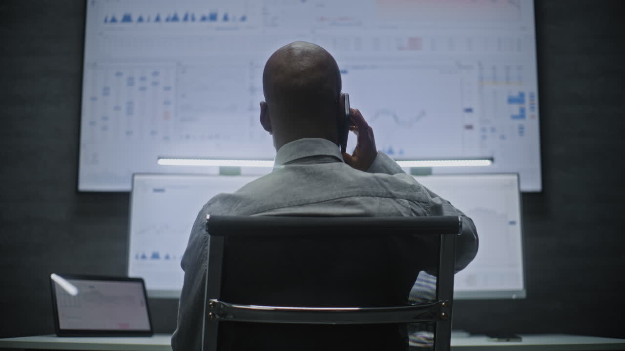 Financial Analyst Talking in Wireless Headphones, Monitoring Exchange Market Charts on Computer with Multi-Monitor Workstation. African American Businessman Works in Office at Night. Vertical Shot.