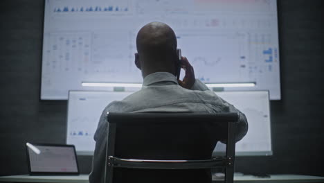 financial analyst talking in wireless headphones, monitoring exchange market charts on computer with multi-monitor workstation. african american businessman works in office at night. vertical shot.
