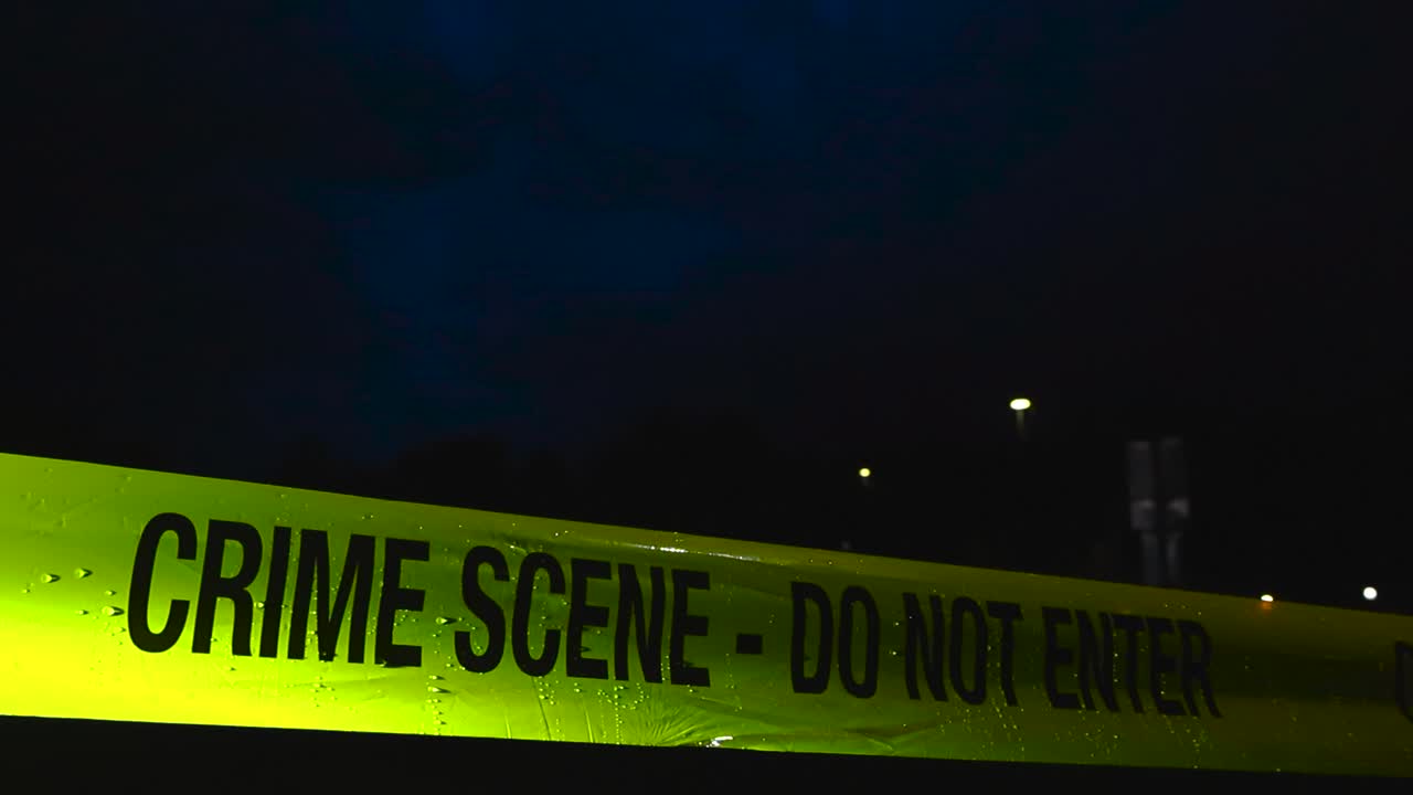 Close up or closeup footage of a yellow colored Crime Scene Do Not Enter police caution tape ribbon pulled in front of a dark blue stormy sky during a rainy evening or night. Water drops visible