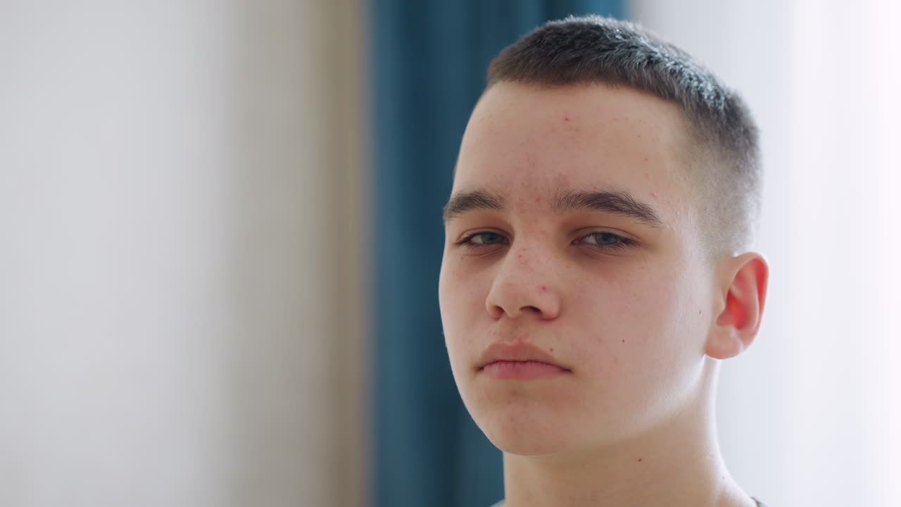 Close up of young student boy with confident and thoughtful expression standing indoors near soft natural light, showing clear skin and short haircut against calm minimal background