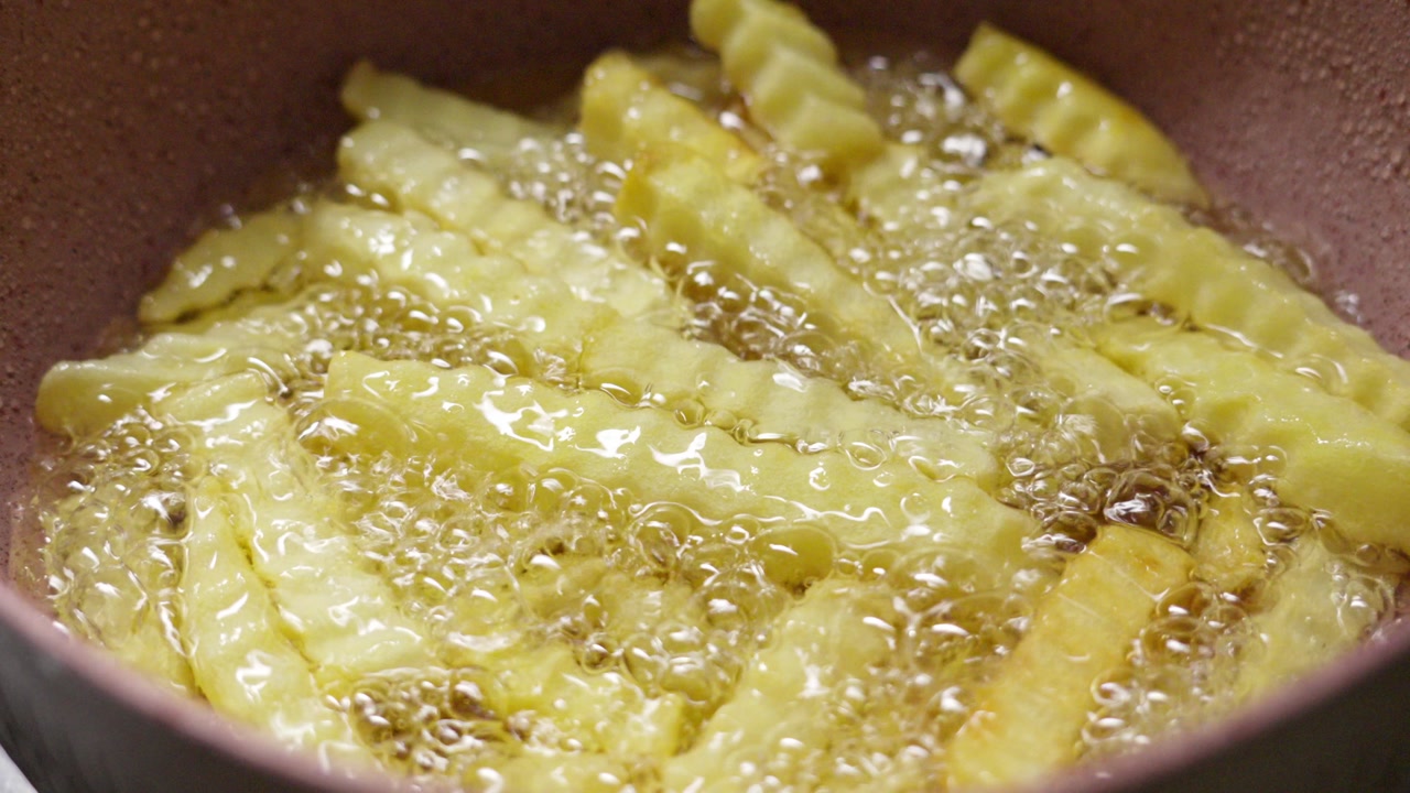 Macro shot of crinkle cut french fries frying in bubbling hot oil. Golden potato cooking in a deep fryer in Indonesia. Delicious fast food preparation for a snack or meal