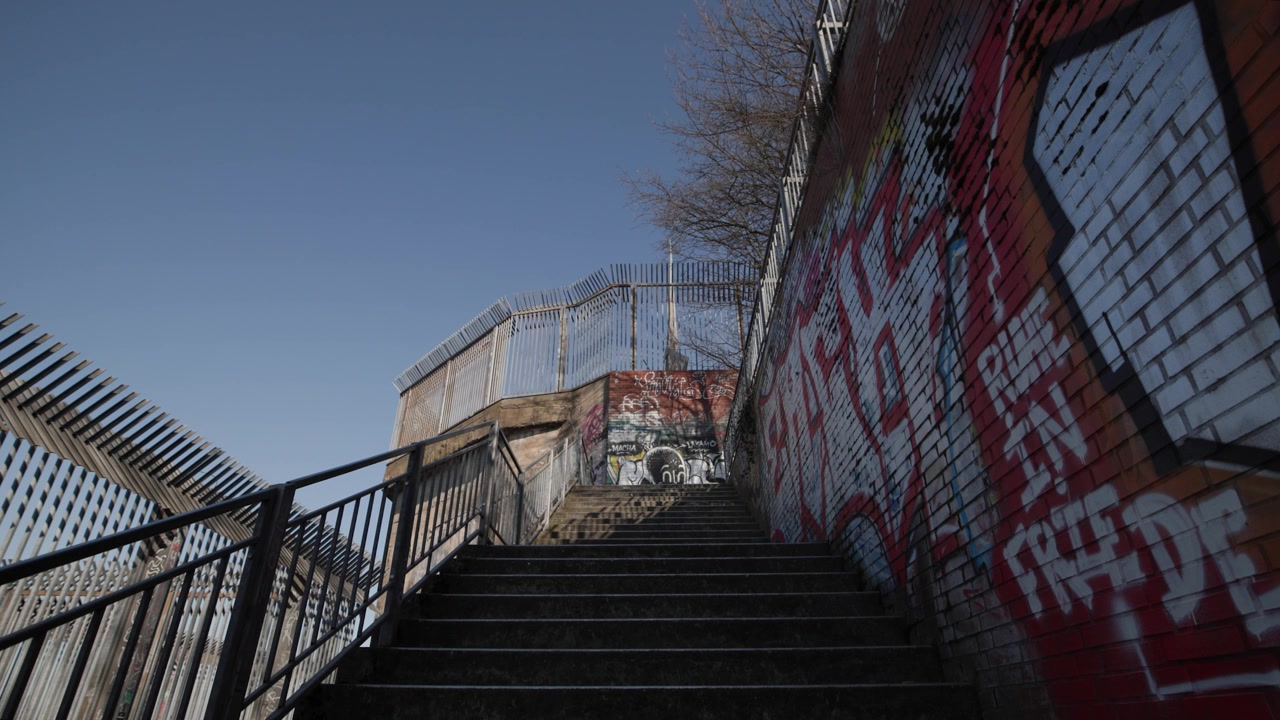 Et lavvinkelbillede, der ser op ad en række udendørs urbane betontrapper, omgivet af metalsikkerhedshegn og murstensvægge dækket af farverig graffitikunst. Klar blå himmel