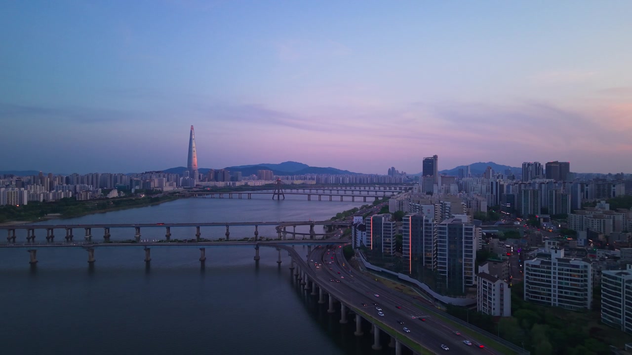 Ripresa aerea in allontanamento sull'autostrada sinuosa Gangbyeonbuk-ro al crepuscolo rivela i ponti del fiume Han e la Lotte World Tower sullo sfondo dello skyline di Seul al tramonto, con sfumature rosa e viola