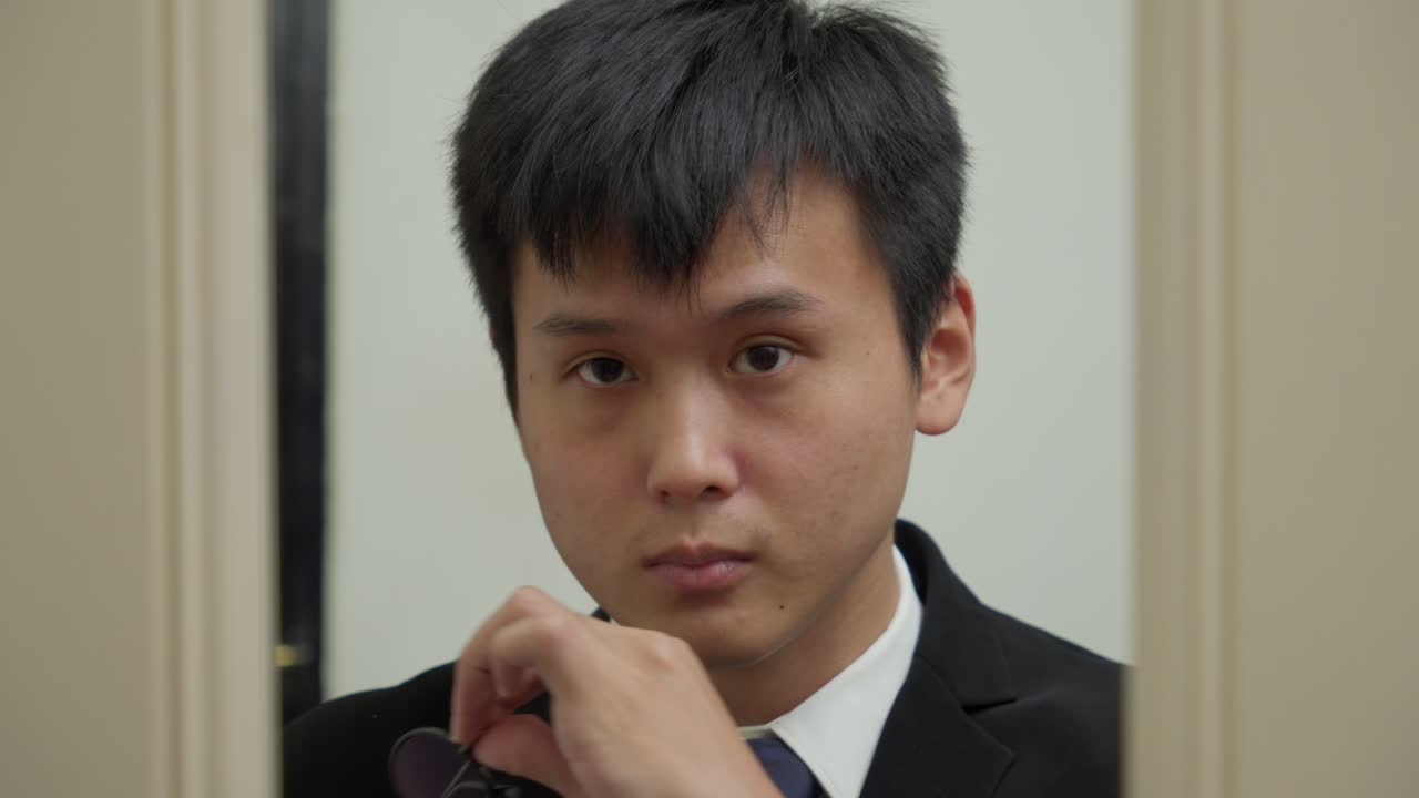 아시아인 남자가 목걸이 타이를 조정하고, 안경을 착용하고, 거울에서 반사를 신중하게 확인합니다.