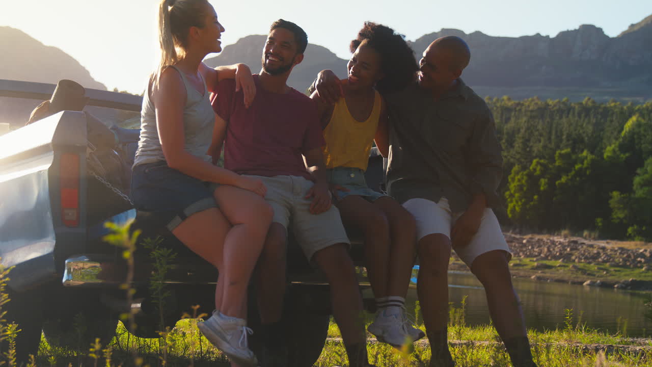 Friends With Backpacks Sitting On Tailgate Of Pick Up Truck On Road Trip By Lake In Countryside