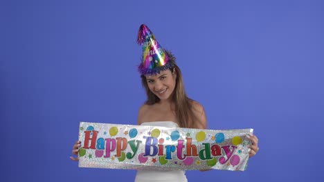 Eine-Junge-Frau-Posiert-Mit-Einem-Happy-Birthday-Schild-Und-Einem-Partyhut,-Isoliert-Auf-Einem-Blauen-Studiohintergrund