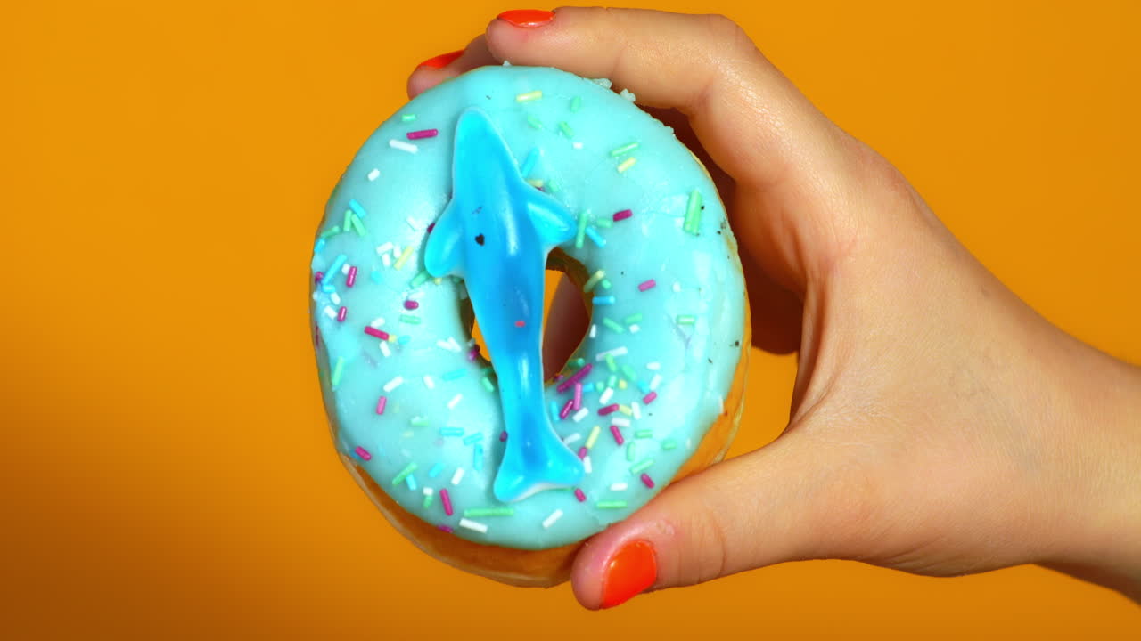 A child’s hand holds a blue frosted donut topped with colorful sprinkles and a gummy shark, set against a vibrant orange background.