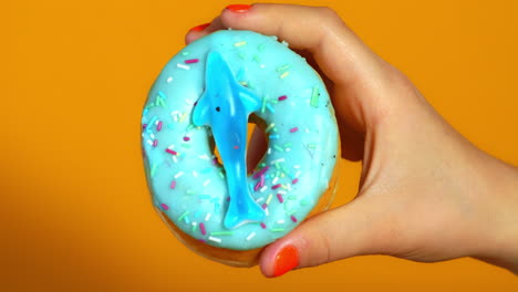 a child’s hand holds a blue frosted donut topped with colorful sprinkles and a gummy shark, set against a vibrant orange background.