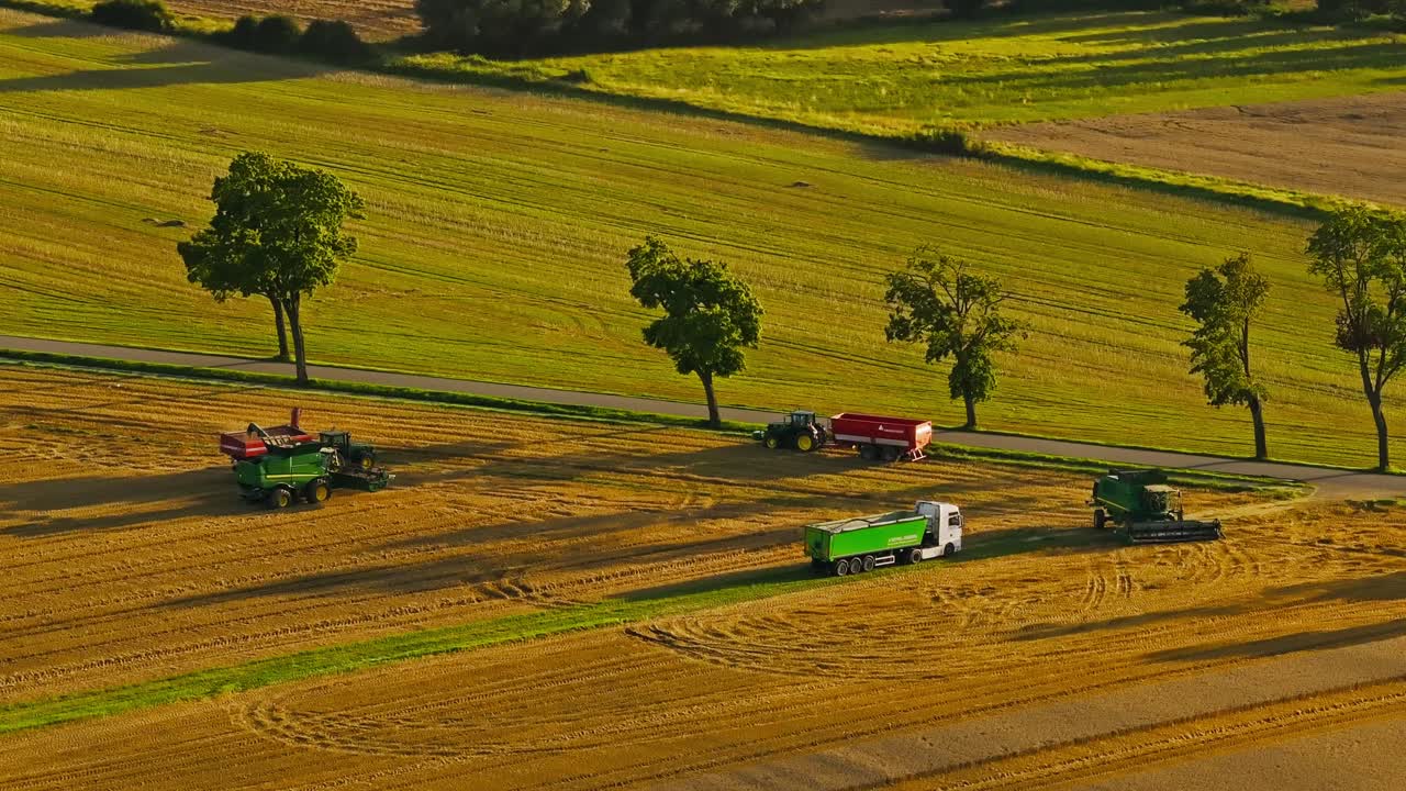 Tractors and trucks collect wheat while drone pans left across rural farmlands