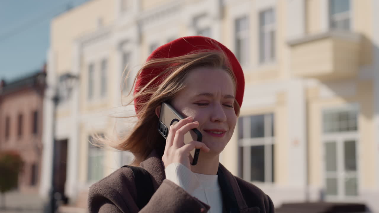 Donna gioiosa con berretto rosso in ambiente urbano. Ritratto all'aperto di donna sorridente con capelli scompigliati dal vento. Scena cittadina casuale con una donna deliziata che indossa un berretto rosso e si gode il clima soleggiato.