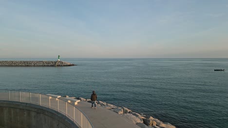 a woman walks alone at the beach dock, seaside of arenys de mar, catalonia aerial drone shot, sightseeing the blue sea, ocean water