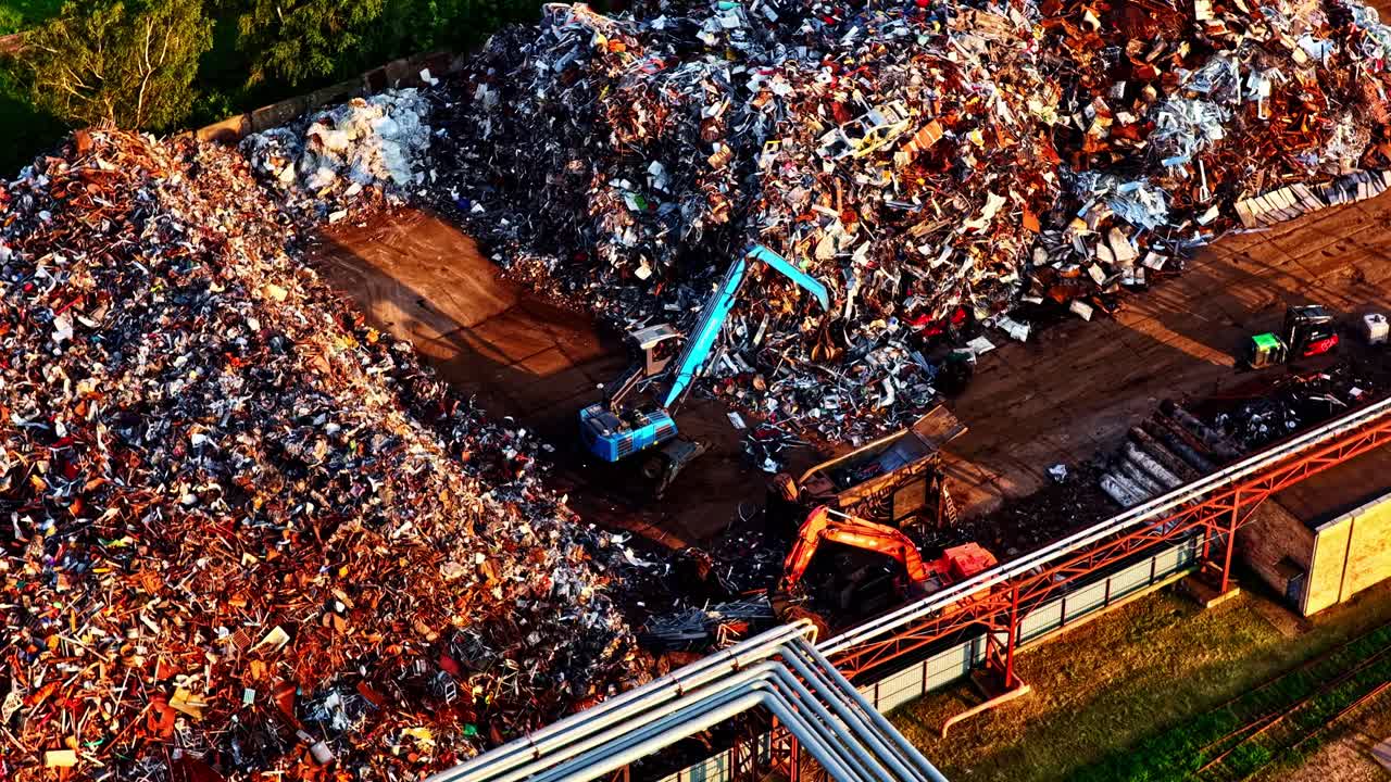 A dramatic aerial parallax shot orbits a large scrap metal yard at sunset, showing excavators sorting through massive piles of industrial waste for recycling