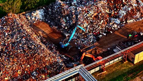 a dramatic aerial parallax shot orbits a large scrap metal yard at sunset, showing excavators sorting through massive piles of industrial waste for recycling