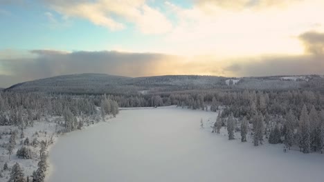 Large-frozen-lake-between-snow-covered-conifers-with-the-high-Harz-mountains-in-Germany-in-the-background,-while-a-thick-cloud-cover-slides-over-the-sun-and-blue-sky