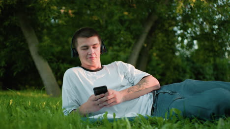trabajador remoto con auriculares acostado en un campo cubierto de hierba, asintiendo con la cabeza a la música mientras opera el teléfono con una sonrisa sutil y un brazo tatuado, rodeado de exuberante vegetación