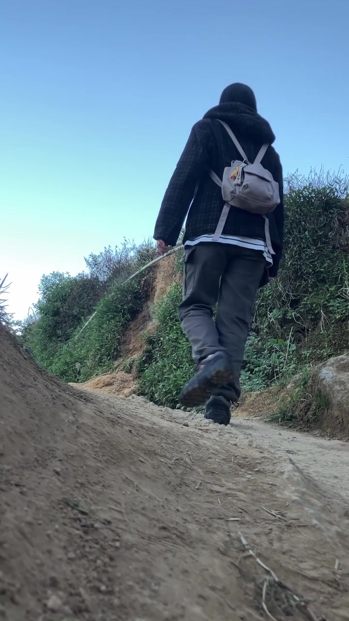 Plano vertical 9:16 de baixo ângulo com vista traseira de uma mulher muçulmana usando hijab, casaco e mochila, a fazer trilha por uma trilha de montanha íngreme de terra contra um céu azul claro.