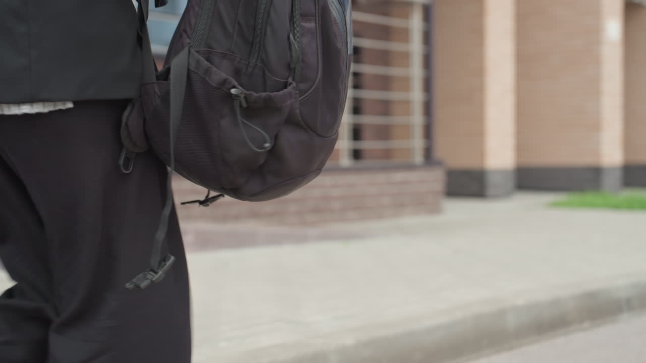 Black backpack on sidewalk during back to school commute, student adjusting straps near brick campus entrance, closeup of zipper and worn fabric, morning routine preparing books and laptop for class