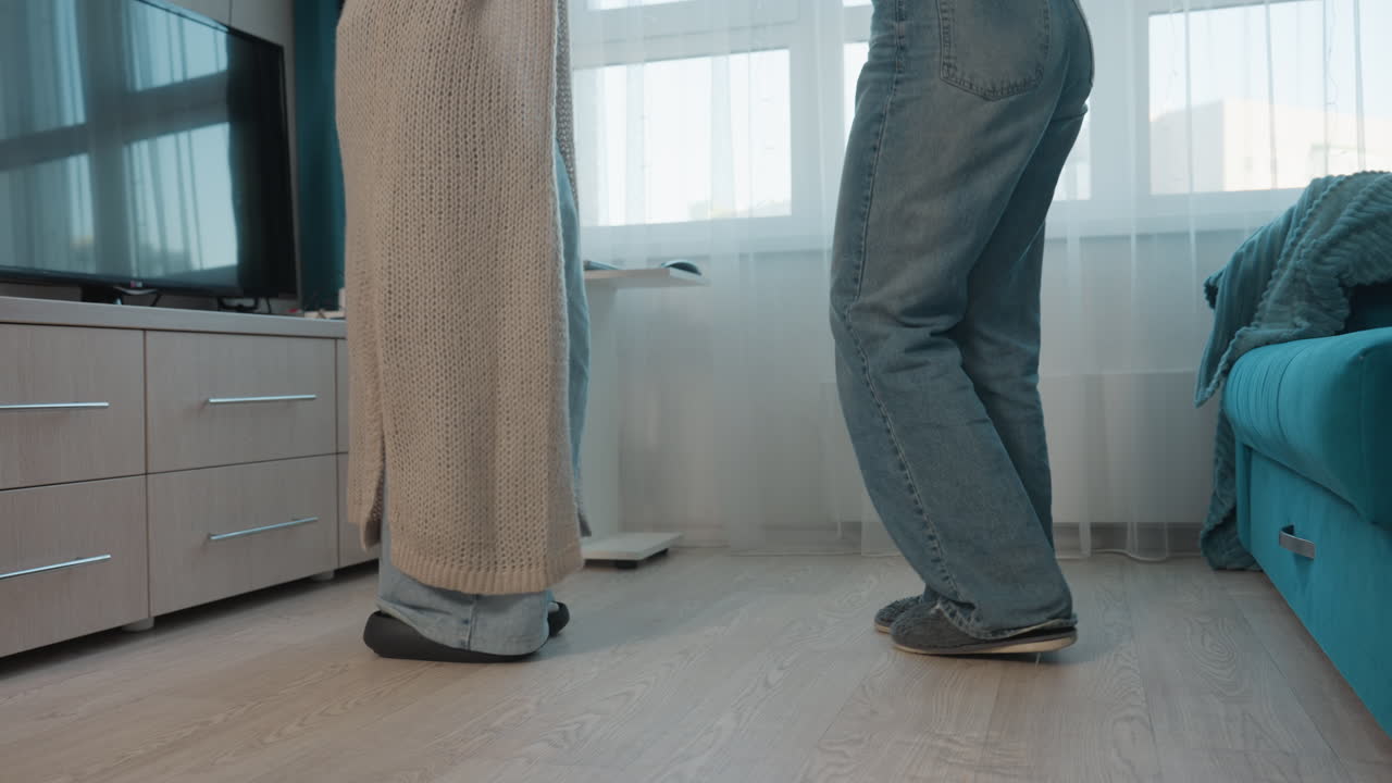 CloseUp Floor Feet Showing Two People Standing In Living Room, Partners Frozen In Awkward Pause Near Sofa, Slippers And Denim Visible, Soft Window Light, Subdued Domestic Tension And Intimate