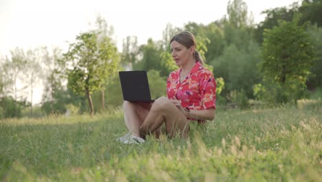 a middle-aged woman sits on the grass in the park. using a video link on a laptop, she conducts a conversation.