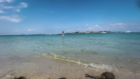 View-of-beautiful-beach-in-Saint-Nicolas-island,-in-background-a-guy-taking-a-ride-on-a-paddle