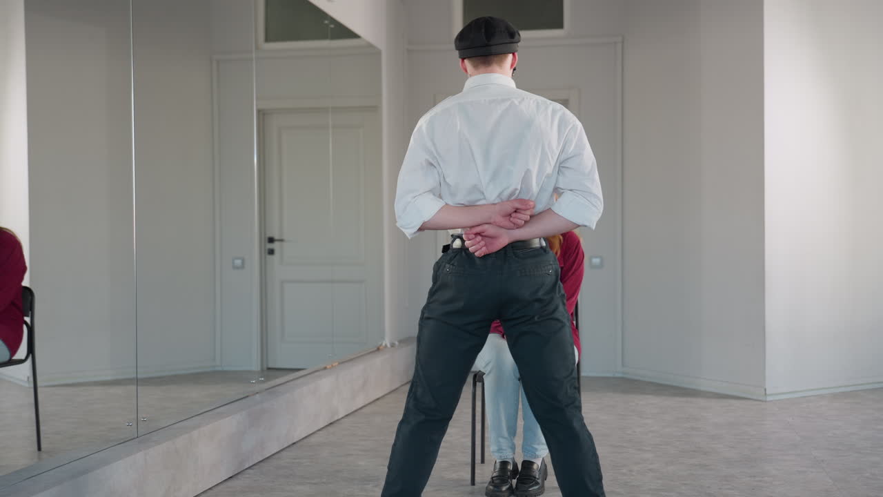 Back view of man in white shirt and black pants extending arms while standing in front of seated woman in red shirt, engaging in playful interaction inside bright modern room with large mirrors