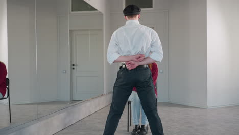back view of man in white shirt and black pants extending arms while standing in front of seated woman in red shirt, engaging in playful interaction inside bright modern room with large mirrors