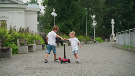 two young siblings joyfully play around a scooter, both with their hands on the handlebar on a paved walkway with electric lamp stand around