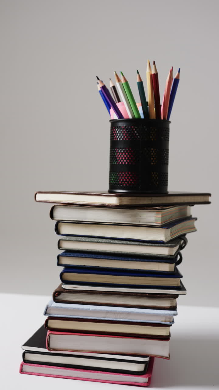 Vertical video of crayons in cup on stack of books on grey background, in slow motion