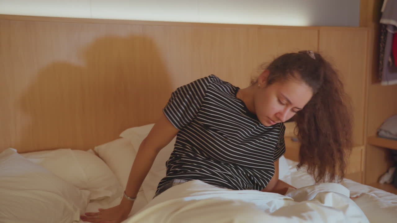 Giovane donna seduta sul letto d'hotel, si strofina gli occhi e si tira indietro i capelli, scruta la stanza e raccoglie i pensieri, testiera in pannelli di legno, valigia visibile vicino ai piedi del letto, espressione determinata, zainista.