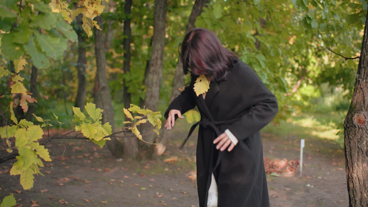 젊은 여성이 공원에서 몸을 숙여 코트 벨트를 묶으며 마른 잎을 턱 근처에 대고, 잎을 사용해 찻잔을 들어 올리는 차분한 가을 풍경이에요. 부드러운 빛과 조용한 숲 바닥이 녹색 캐노피 아래 펼쳐져요.