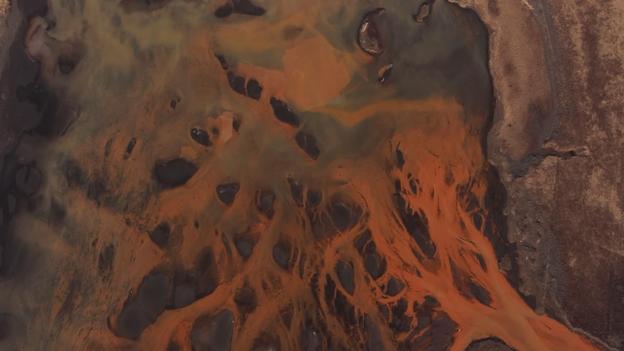 Abstract aerial view of the Þjórsá River in Iceland, where volcanic sediment creates vivid orange streams over dark terrain, forming a surreal natural pattern.