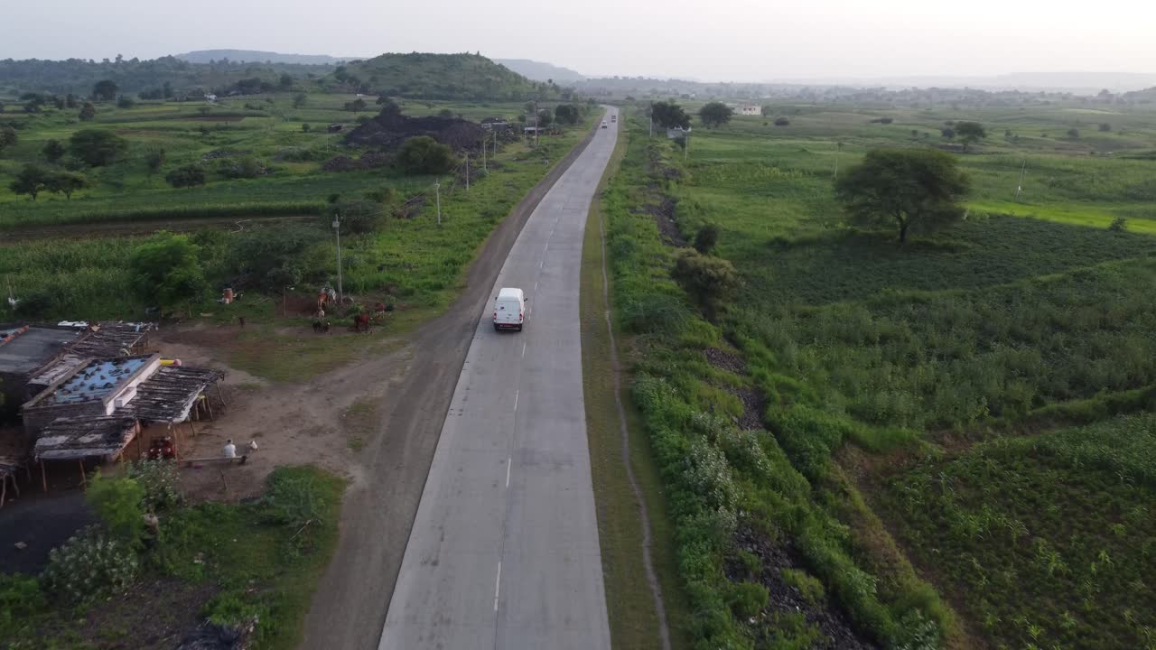 Drone following a goods carrier van traveling on a rural Indian road of good quality with the surroundings of green farmlands and village homes