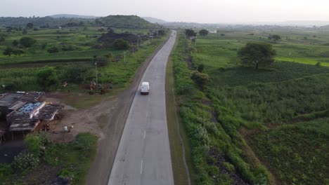 Drone-Siguiendo-Una-Furgoneta-De-Transporte-De-Mercancías-Que-Viaja-Por-Una-Carretera-India-Rural-De-Buena-Calidad-Con-Los-Alrededores-De-Tierras-De-Cultivo-Verdes-Y-Casas-De-Aldea