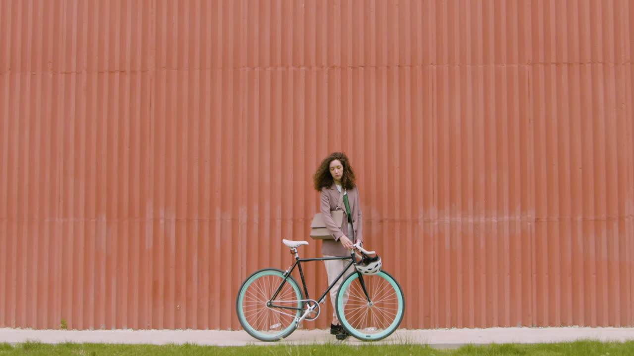 정장 차림의 젊은 여성이 자전거와 함께 걷고 있다가 멈춰 서서 조립식 금속 건물 앞에서 카메라를 바라보고 있습니다.