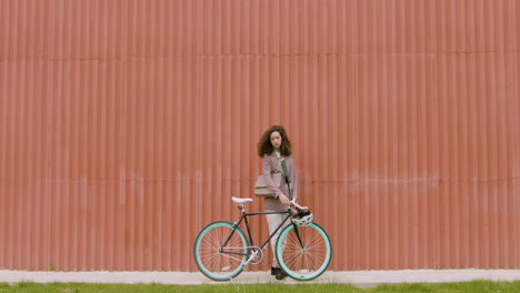 mujer joven con ropa formal caminando con su bicicleta, luego deteniéndose y mirando la cámara frente a un edificio de metal prefabricado