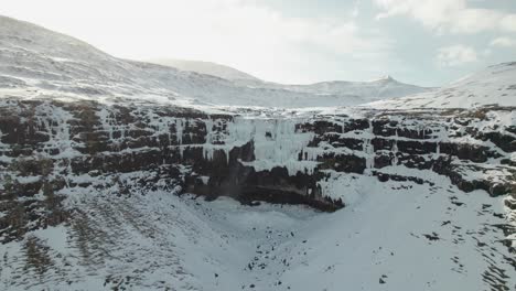 4k aerial drone shot of 140 meter tall frozen waterfall fossurin i fossá, the largest waterfall on the faroe islands on streymoy on a cold and sunny winter day in a landscape covered in snow