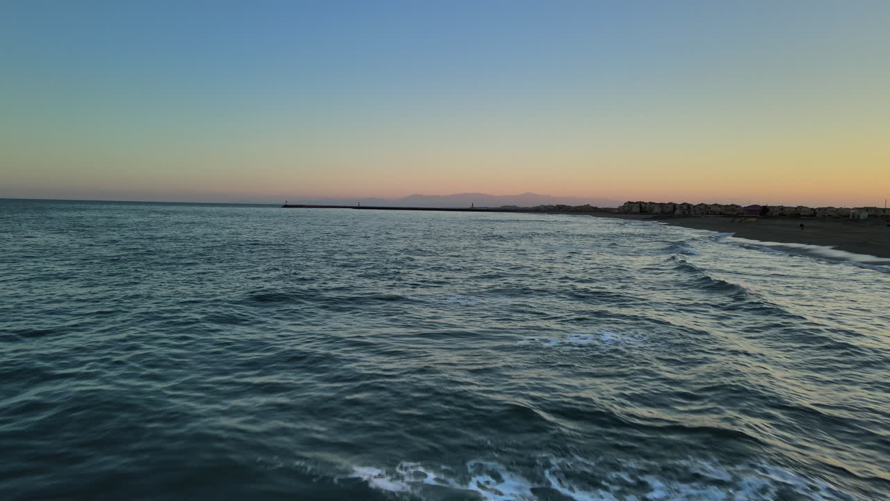 Onde lambiscono dolcemente la riva di una spiaggia tranquilla. Il sole tramonta all'orizzonte, proiettando colori caldi nel cielo e creando un'atmosfera serena