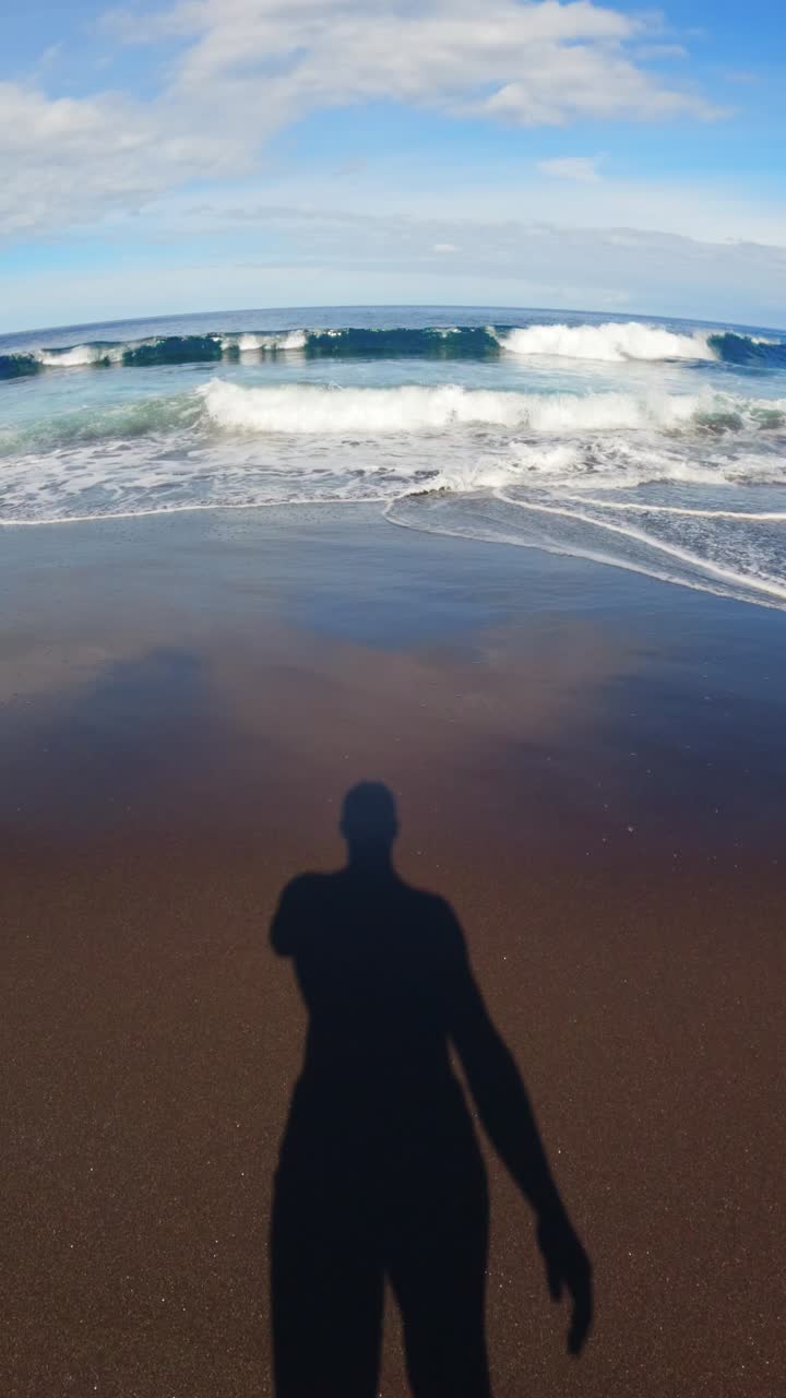 POV man's feet and his shadow walking on a dark sand beach towards the ocean with waves