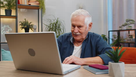 happy senior old man grandfather sitting on sofa at table working on laptop pc at office home room