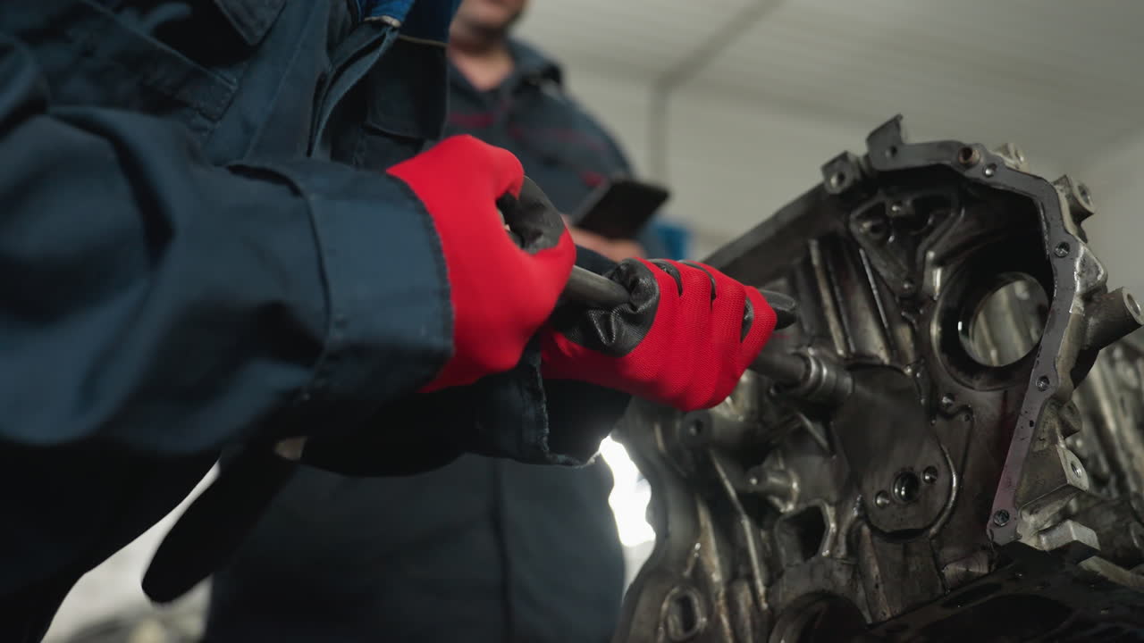 primer plano de un ingeniero con un pendiente magnético usando una llave inglesa para aflojar una nuez en un motor, con guantes rojos, mientras su colega observa el proceso en el taller