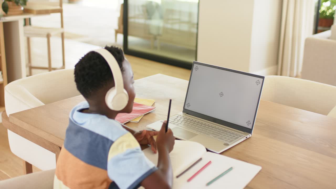 Putting on headphones at desk, child is glancing at laptop while jotting notes awaiting content