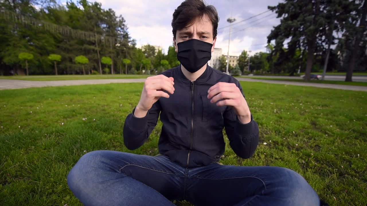Happy young man removing a black protective mask from his face and looking at the camera while resting on the grass in park. Driving cars on background at sunset. Quarantine and Corona Virus idea