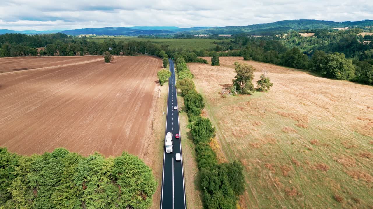 Sobrevoo de estrada reta com carros através de campos agrícolas e floresta